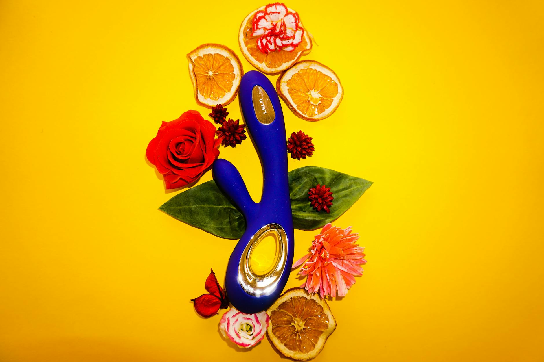 Mano extendida sosteniendo un vibrador personal azul rodeado de flores y naranjas sobre fondo amarillo